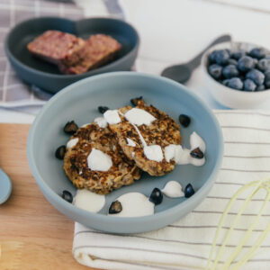 Blue silicone suction plate with banana pancakes on and yoghurt and blueberries.