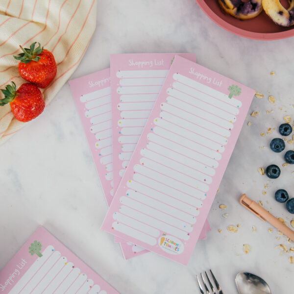 three shopping list pads on marble background surrounded by fruit, a pen and some baby cutlery